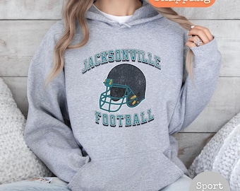 Op de afbeelding: Een grijs hoodie met de tekst "JACKSONVILLE FOOTBALL" en een vintage-stijl voetbalhelm afbeelding.