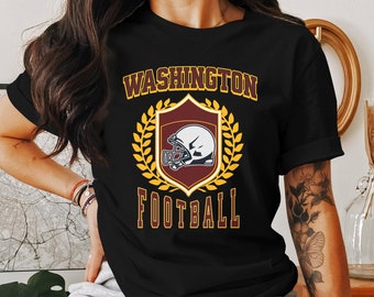 Op de afbeelding: Zwart T-shirt met een gouden en kastanjebruine afbeelding van een voetbalhelm in een schild met de tekst "WASHINGTON FOOTBALL" erboven en eronder.