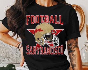 May include: Black t-shirt featuring a red star with the words "FOOTBALL SAN FRANCISCO" in red. A gold football helmet with a red lightning bolt is positioned in the centre of the star.