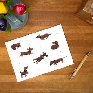 May include: A white card with illustrations of brown dachshunds in different poses. The card is on a wooden surface, with a pen, a vase of tulips, and a wooden desk organiser.