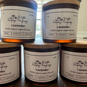 Puede incluir: Cuatro tarros de velas de vidrio ámbar con tapas de madera. Cada tarro tiene una etiqueta blanca con el texto "Simply Foraging" y "Lavender" en negro. La etiqueta también indica "100% Organic Hand-Poured" y enumera los ingredientes: cera de abejas, aceite de coco y aceite esencial.