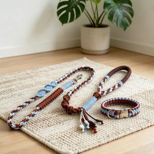 Puede incluir: Un conjunto de correa y collar trenzados para mascota con una paleta de colores azul claro, blanco y marrón oscuro. La correa incluye un mosquetón giratorio de metal, mientras que el collar cuenta con una hebilla de metal plateado.
