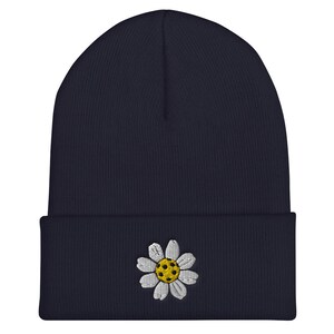 May include: A navy blue beanie with a white daisy embroidered on the front. The daisy has a yellow center with white petals.