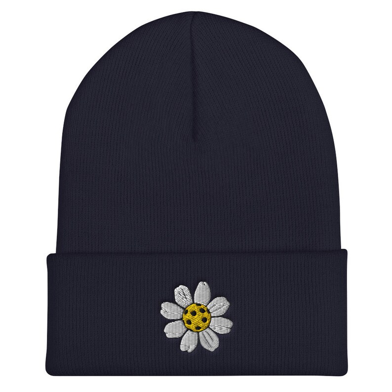 May include: A navy blue beanie with a white daisy embroidered on the front. The daisy has a yellow center with white petals.