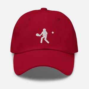Puede incluir: Una gorra de b&eacute;isbol roja con un dise&ntilde;o bordado en blanco de una persona jugando al pickleball.