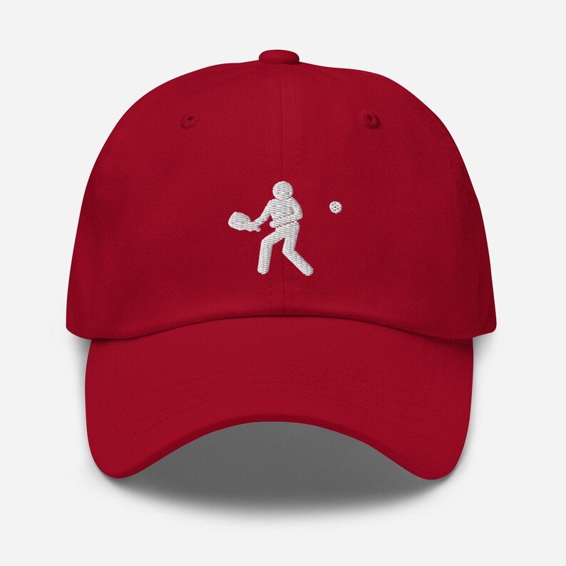 Puede incluir: Una gorra de b&eacute;isbol roja con un dise&ntilde;o bordado en blanco de una persona jugando al pickleball.