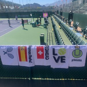 May include: Five white towels with different designs hanging on a green fence. The towels feature a tennis racket, the Spanish flag, a Swiss flag, the word "Fedhead", the word "Love" with a tennis ball, and a tennis ball with a crown and the word "princess".