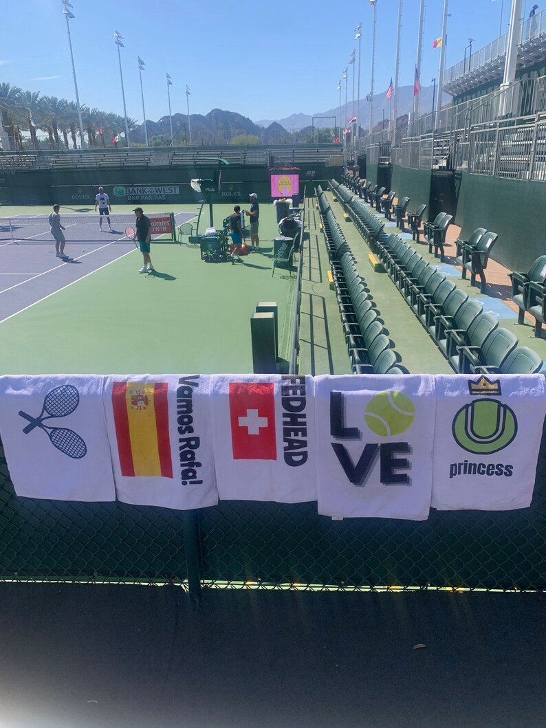 May include: Five white towels with different designs hanging on a green fence. The towels feature a tennis racket, the Spanish flag, a Swiss flag, the word "Fedhead", the word "Love" with a tennis ball, and a tennis ball with a crown and the word "princess".