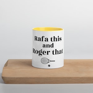 May include: White ceramic mug with a yellow interior, featuring the text "Rafa this and Roger that" in black. A tennis racket and ball graphic are below the text. The mug is on a wooden surface.
