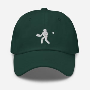 Puede incluir: Una gorra de b&eacute;isbol verde oscuro con una imagen bordada en blanco de una persona jugando un deporte con una raqueta y una pelota.