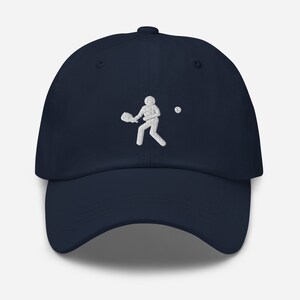 Puede incluir: Una gorra de b&eacute;isbol azul marino con un dise&ntilde;o bordado en blanco de una persona jugando al tenis.