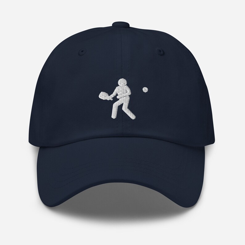 Puede incluir: Una gorra de b&eacute;isbol azul marino con un dise&ntilde;o bordado en blanco de una persona jugando al tenis.