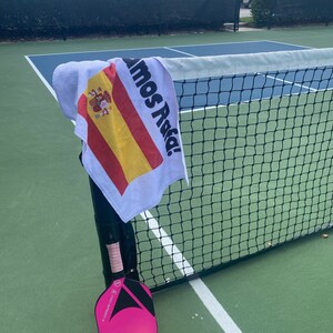 May include: A pink and black pickleball paddle with a white grip and a yellow pickleball on a green court. A white towel with the Spanish flag and the text "Vamos Asia!" is hanging on a black net.