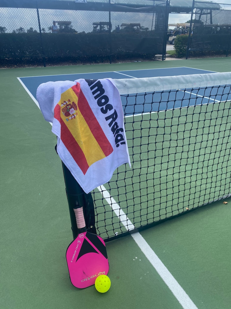 May include: A pink and black pickleball paddle with a white grip and a yellow pickleball on a green court. A white towel with the Spanish flag and the text "Vamos Asia!" is hanging on a black net.