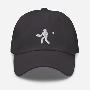 Puede incluir: Una gorra de b&eacute;isbol gris oscuro con un dise&ntilde;o bordado en blanco de una persona jugando un deporte con una raqueta y una pelota.