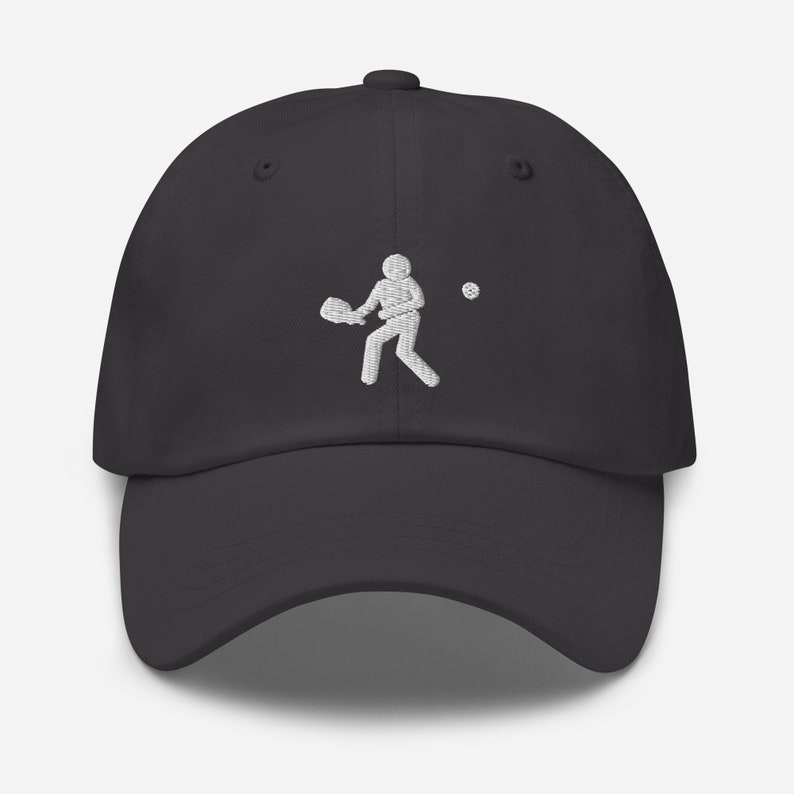Puede incluir: Una gorra de b&eacute;isbol gris oscuro con un dise&ntilde;o bordado en blanco de una persona jugando un deporte con una raqueta y una pelota.