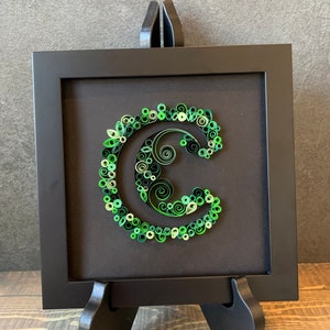 Può includere: Un'opera d'arte in carta incorniciata che presenta la lettera "C" realizzata con riccioli di carta verde. L'opera d'arte è esposta su un cavalletto nero.