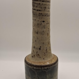 Może przedstawiać: Wysoki, ceramiczny wazon o cylindrycznym kształcie. Wazon ma nakrapiane, jasnobeżowe ciało z ciemnobrązowymi akcentami i ciemnoniebiesko-zieloną podstawą. Wazon ma teksturowaną powierzchnię i rozszerzoną podstawę.