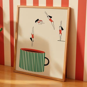 May include: A framed art print featuring a whimsical illustration. The artwork depicts stylized figures in red swimsuits diving, along with a large green and white striped coffee cup. The frame is made of light-colored wood.