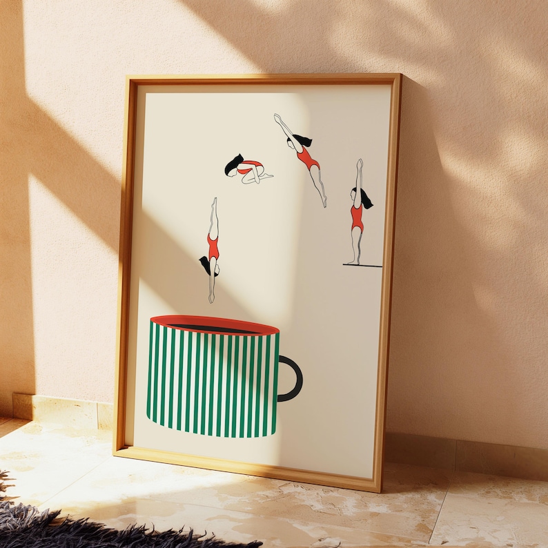 May include: A framed art print featuring a whimsical design. The artwork depicts four figures in red swimsuits diving, set against a cream background. A large coffee cup with green and white stripes and a red rim is in the foreground.