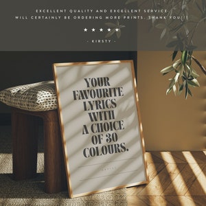 May include: A framed print with the text "YOUR FAVOURITE LYRICS WITH A CHOICE OF 30 COLOURS." The print has a gold frame and is displayed on a wooden bench. The background includes a pillow and a wall with sunlight streaming through blinds.