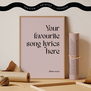 May include: A framed print with the text "Your favourite song lyrics here" in a stylish font. A rolled-up print tied with twine and a spool of twine are next to the framed print. The background is a light pink color.