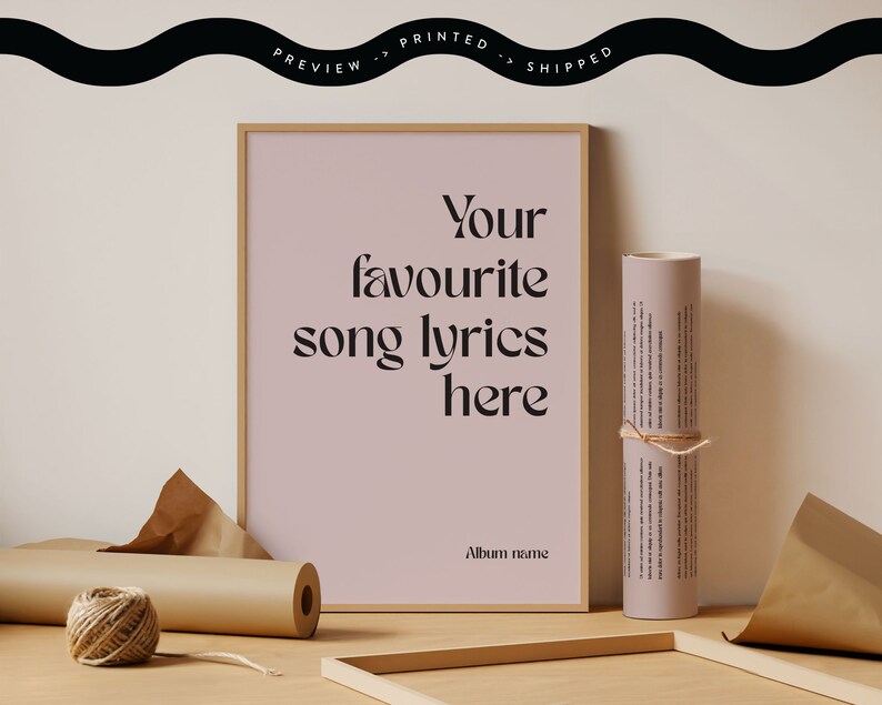 May include: A framed print with the text "Your favourite song lyrics here" in a stylish font. A rolled-up print tied with twine and a spool of twine are next to the framed print. The background is a light pink color.