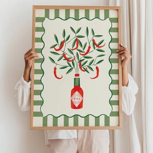 May include: A framed art print featuring a stylized illustration of a chili pepper plant. The artwork depicts red chili peppers growing from a bottle of hot sauce, set against a cream background with a green and white striped border. The frame is made of wood.