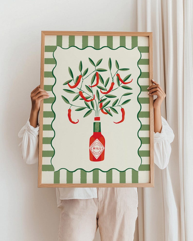 May include: A framed art print featuring a stylized illustration of a chili pepper plant. The artwork depicts red chili peppers growing from a bottle of hot sauce, set against a cream background with a green and white striped border. The frame is made of wood.