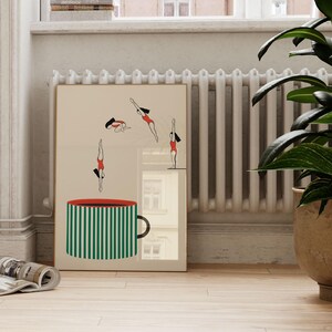 May include: A framed art print featuring a large green and white striped mug with a red rim. Above the mug, stylized figures in red swimsuits are depicted diving. The artwork is set against a neutral background.