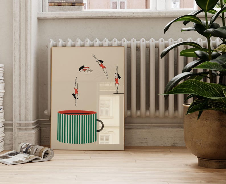 May include: A framed art print featuring a large green and white striped mug with a red rim. Above the mug, stylized figures in red swimsuits are depicted diving. The artwork is set against a neutral background.