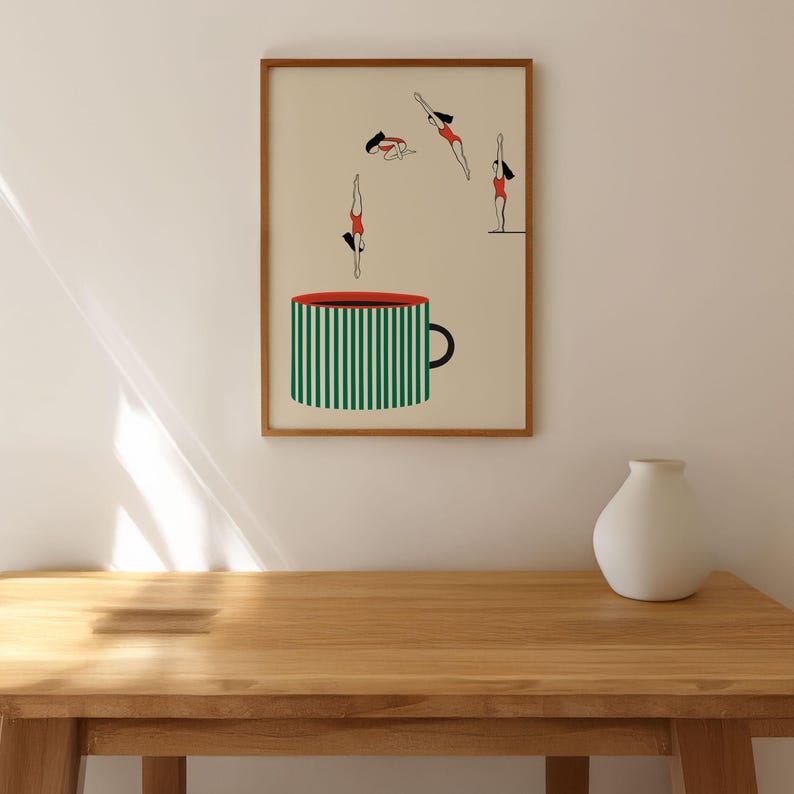 May include: A framed art print featuring a large coffee mug with green and white stripes, and four figures in red swimsuits diving into the mug. The artwork is set against a beige background.