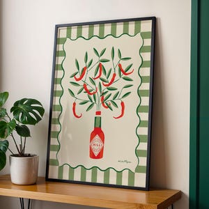 May include: Framed art print featuring a stylized illustration of red chili peppers growing from a Tabasco sauce bottle. The artwork has a cream background with a green and white striped border and a black frame. A potted plant sits to the left.