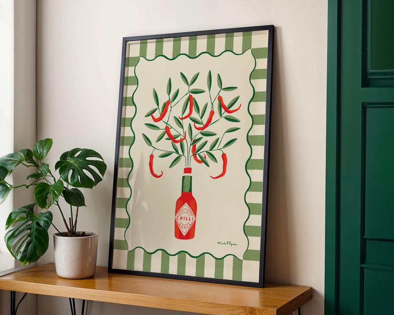 May include: Framed art print featuring a stylized illustration of red chili peppers growing from a Tabasco sauce bottle. The artwork has a cream background with a green and white striped border and a black frame. A potted plant sits to the left.