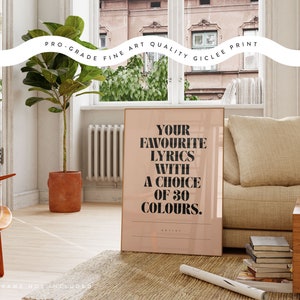 May include: A framed print with the text "YOUR FAVOURITE LYRICS WITH A CHOICE OF 30 COLOURS" in black on a light pink background. The print is displayed in a room with a sofa, wooden chair, and a potted plant.