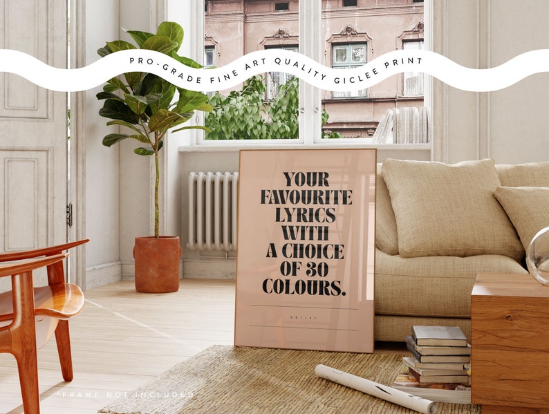 May include: A framed print with the text "YOUR FAVOURITE LYRICS WITH A CHOICE OF 30 COLOURS" in black on a light pink background. The print is displayed in a room with a sofa, wooden chair, and a potted plant.
