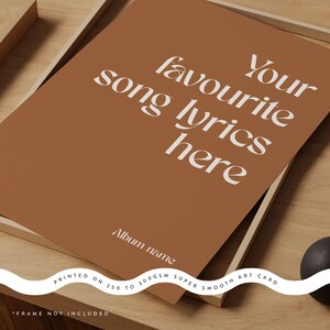 May include: A framed art print with a warm brown background. The print features the text "Your favourite song lyrics here" in a white, stylized font. Below, it reads "Album name." The bottom of the print states "PRINTED ON 250 TO 300GSM SUPER SMOOTH ART CARD."