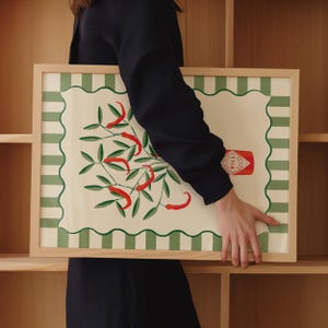 May include: A framed art print featuring a botanical illustration of red chili peppers and green leaves. The artwork has a cream-colored background with a green and white striped border and a wooden frame. A red chili sauce bottle is also depicted.