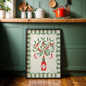 May include: A framed art print featuring a stylized illustration of chili peppers. The artwork depicts red chili peppers growing from a bottle of chili sauce, set against a cream background with a green and white striped border. The frame is black.