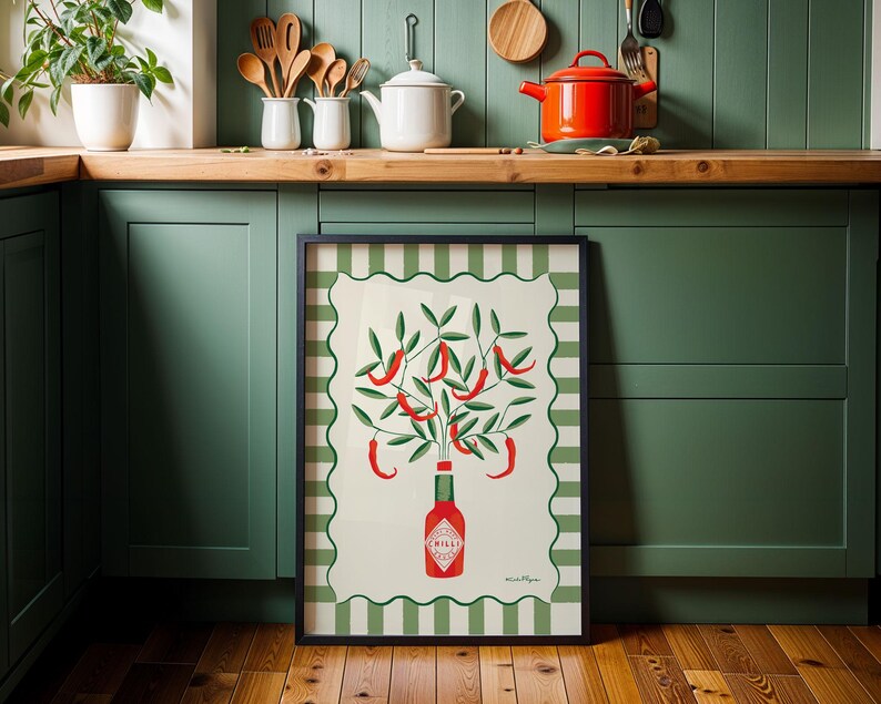 May include: A framed art print featuring a stylized illustration of chili peppers. The artwork depicts red chili peppers growing from a bottle of chili sauce, set against a cream background with a green and white striped border. The frame is black.