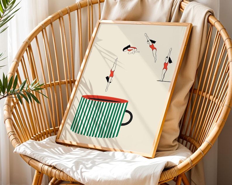 May include: A framed art print featuring a whimsical illustration of divers in red swimsuits and a large green and white striped coffee mug. The artwork is displayed on a light beige background and is set in a natural wood frame.