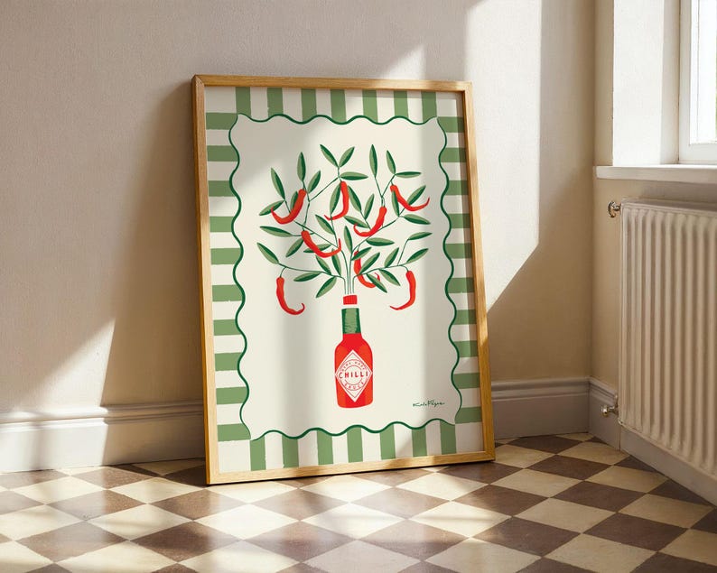 May include: A framed art print featuring a stylized illustration of red chili peppers growing from a green bottle labeled "Chilli". The artwork has a cream background with a green and white border, and is set in a wooden frame.
