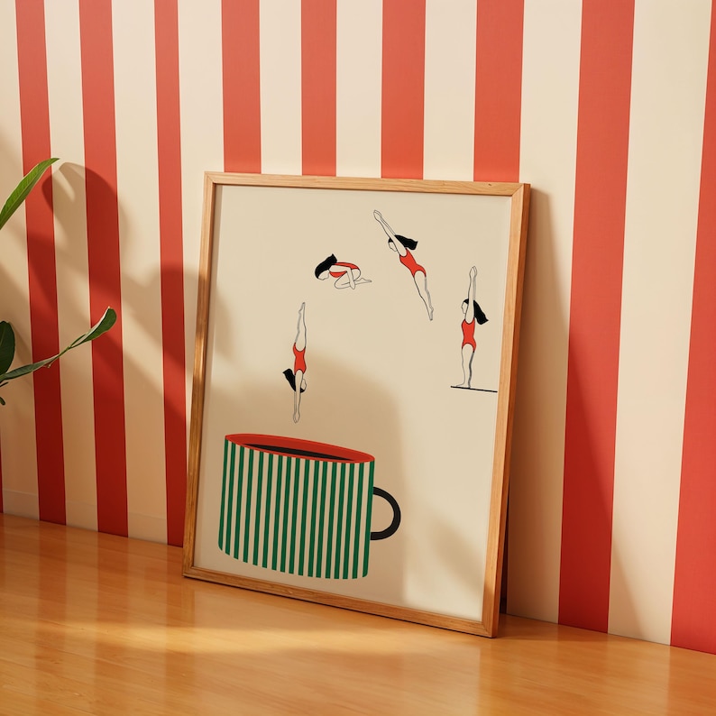 May include: A framed art print featuring a whimsical illustration. The artwork depicts stylized figures in red swimsuits diving, along with a large green and white striped coffee cup. The frame is made of light-colored wood.