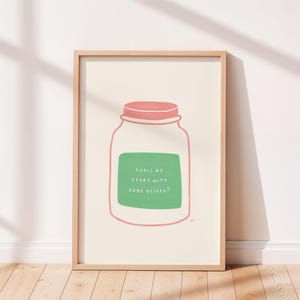 May include: A framed wall art print featuring a line drawing of a glass jar with a pink lid. Inside the jar is a green rectangular label with the white text "SHALL WE START WITH SOME OLIVES?" The print is set against a cream-coloured background.