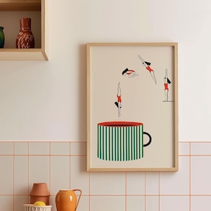 May include: A framed art print featuring a whimsical design. The artwork depicts stylized figures in red swimsuits diving into a large green and white striped coffee cup. The cup has a red rim and a black handle. The background is a light beige.