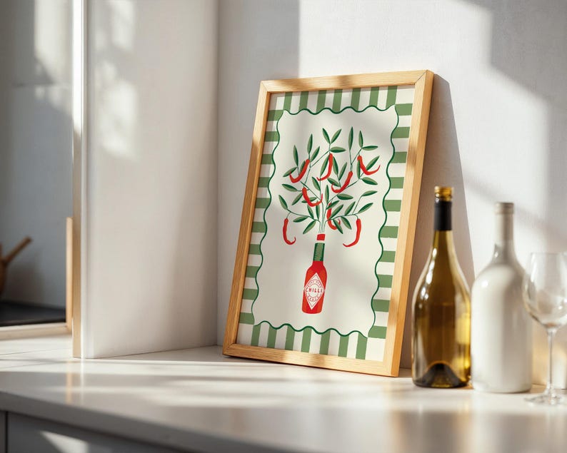 May include: A framed art print featuring a stylized illustration of a Tabasco sauce bottle with chili peppers and green leaves. The print has a green and white striped border and a natural wood frame. A wine bottle and glass are next to the frame.