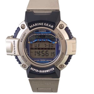 May include: A silver and navy Casio Marine Gear digital watch. The watch face displays the time, date, and depth-barometer readings. The watch has buttons for sensor, adjust, mode, light, and start-stop functions. The watch is water resistant to 100 metres.