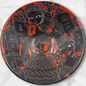 Puede incluir: Un plato de cerámica negro con un diseño dibujado a mano que presenta a dos mujeres y edificios. El diseño está delineado en blanco, con detalles en rojo alrededor del borde y las figuras. El plato tiene un acabado brillante.