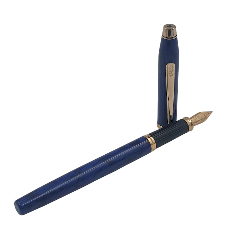 K&ouml;nnte beinhalten: Ein blau marmoriertes F&uuml;llfederhalter mit goldfarbenen Akzenten und einer goldenen Feder. Die Kappe ist abgenommen und neben dem Stift platziert. Auf dem Clip des Stifts steht das Wort "CROSS".