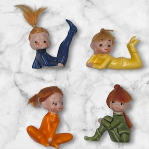 Op de afbeelding: Vier keramische beeldjes van kinderen in verschillende yogahoudingen. Elk beeldje heeft een geschilderd gezicht, rood haar en draagt een andere kleur outfit: blauw, geel, oranje en groen. De beeldjes staan op een wit en grijs marmerpatroon.